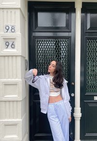 Γκρι φούτερ με φερμουάρ πάνω από λευκή κοντή μπλούζα, μπλε και λευκές ριγέ παντελόνες, κρατώντας γυαλιά ηλίου, leaning πάνω σε μια υφασμάτινη μαύρη πόρτα με αριθμητικές λεπτομέρειες.