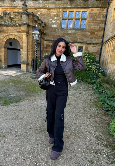 Chaqueta corta de cuero sintético marrón con cuello de borrego blanco, suéter negro de cuello alto, pantalones cargo negros y botas de tobillo marrones. Bolso negro.