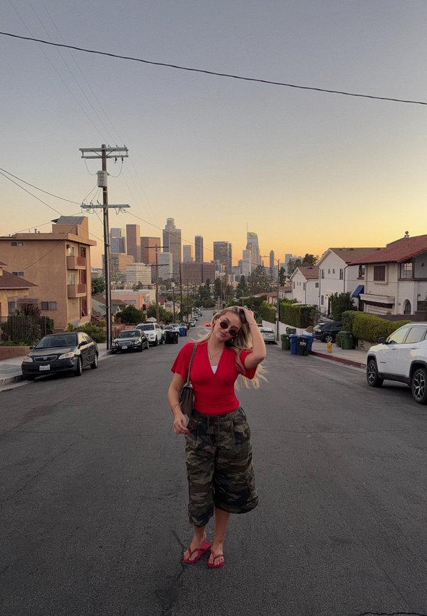 Una mujer con una camiseta roja y pantalones de camuflaje posa en una calle residencial, con coches aparcados y el perfil urbano de Los Ángeles al atardecer.