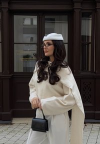 Biała oversize'owa sweter z rękawami w stylu peleryny, czarna torebka, biała beretka i okulary, stojąca na tle ciemnego drewnianego tła.