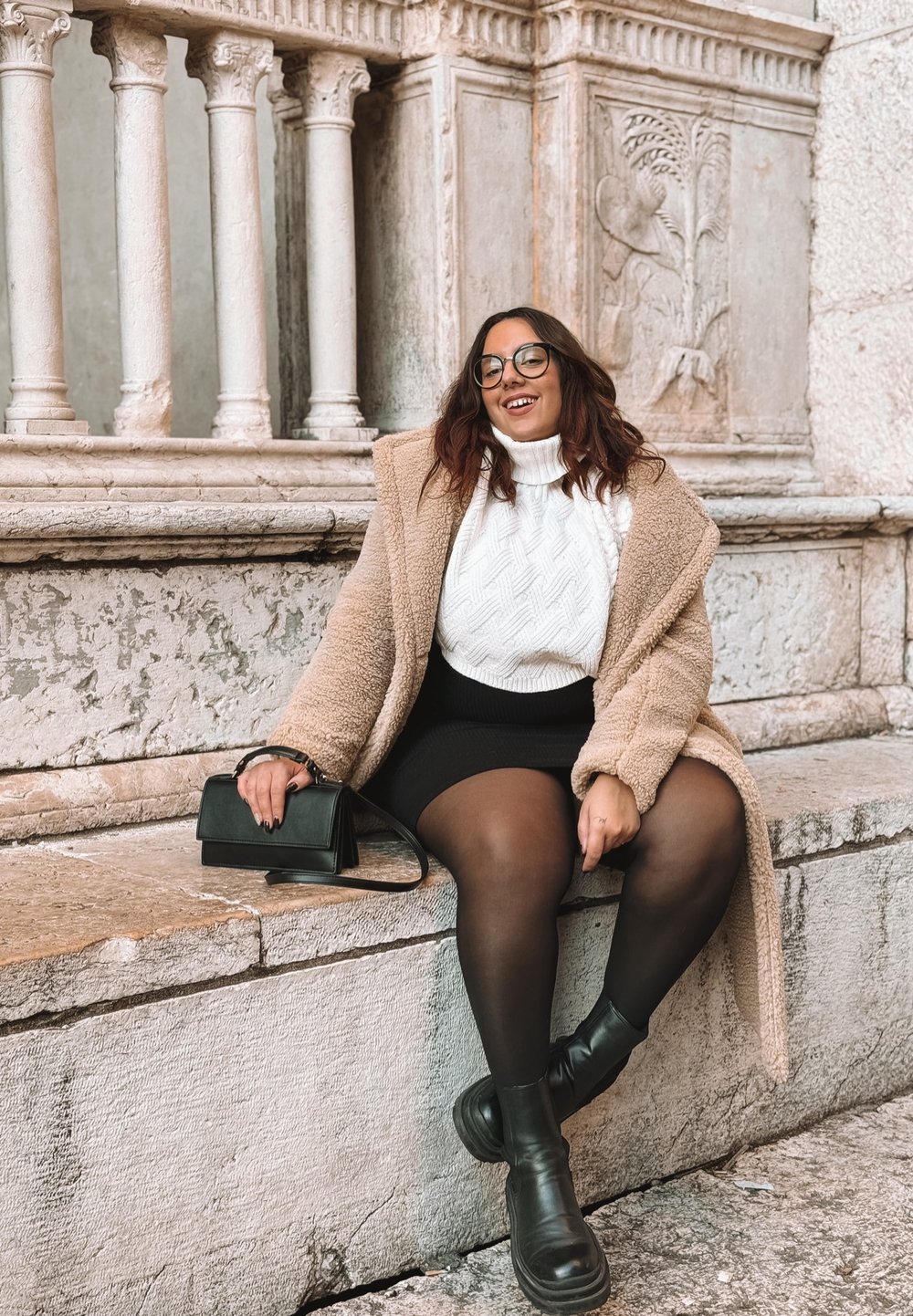 Mulher com óculos vestindo um casaco bege, suéter branco, saia preta e botas, sentada numa borda de pedra, com colunas ornamentadas ao fundo.