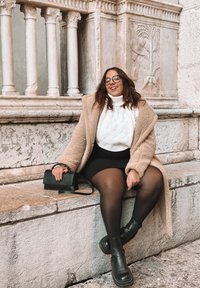 Mulher com óculos vestindo um casaco bege, suéter branco, saia preta e botas, sentada numa borda de pedra, com colunas ornamentadas ao fundo.
