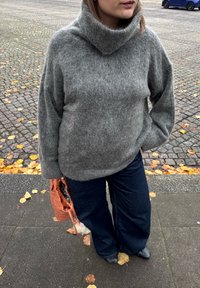 Pulover gri tricotat cu o gâtă înaltă, asociat cu blugi evazați de culoare bleu închis; ține o geantă texturată portocalie.