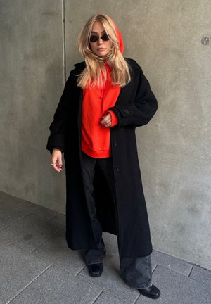 Largo abrigo negro sobre una sudadera roja con capucha, vaqueros negros anchos y botas negras de punta cuadrada; gafas de sol redondas; cabello rubio liso.