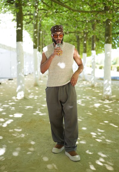 Top sin mangas beige de punto, pantalones grises holgados y zapatos slip-on blancos con acentos marrones, sosteniendo una bebida bajo árboles con hojas.