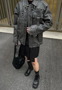 Giacca di pelle grigia con più tasche, cerniere e dettagli in metallo. Abbinata a un vestito nero e stivali neri fino al ginocchio.