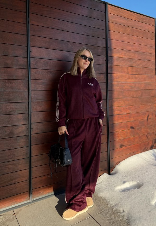 Femme portant des lunettes de soleil et un survêtement bordeaux, debout contre un mur en bois à côté de la neige, tenant un sac à main noir dans une main.