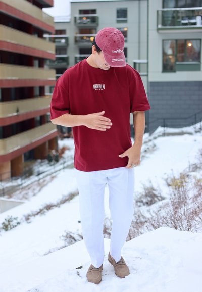 Hombre con gorra roja y camiseta, pantalones blancos y mocasines marrones, de pie en una azotea nevada con edificios urbanos de fondo.