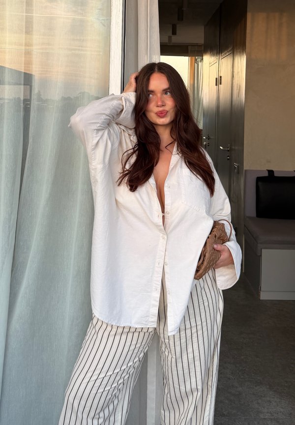Donna con i capelli lunghi e scuri in piedi all'interno vicino a una tenda trasparente, indossa una camicia bianca oversize, pantaloni a righe e tiene una piccola borsa marrone.