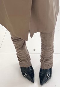 Beige ribbed leg warmers over black pointed-toe shoes with double silver buckles, worn with a beige skirt or coat on a white tiled floor.