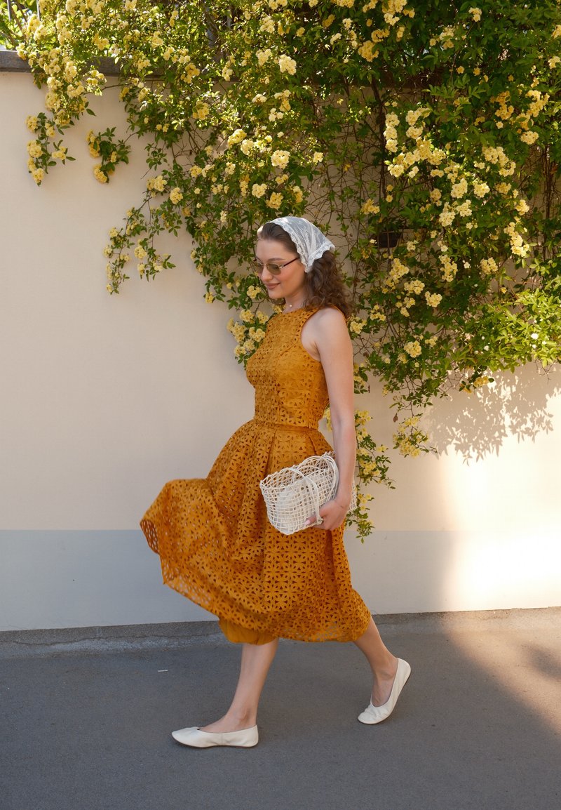 Femme en robe jaune moutarde marchant devant un mur avec des vignes florales jaunes, tenant un sac à main blanc tissé, portant des lunettes de soleil et des chaussures plates blanches.