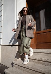 Plastev obleka z karirastim blazerjem, rjavim zgornjim delom, olivno satensko krilo in belimi superge z zelenimi poudarki, se spušča po kamnitih stopnicah.