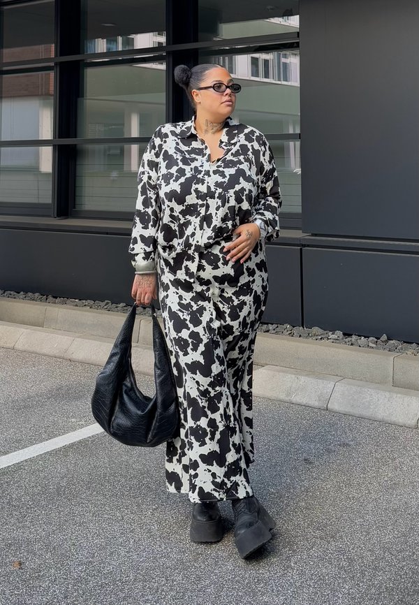 Oversized shirt dress with a black and white cow print, featuring long sleeves, styled with a slouchy black handbag and chunky black shoes.