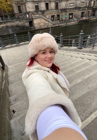 Ένα ανοιχτό μπεζ ψεύτικο γούνινο καπέλο και ένα κρεμ ολόσωμο παλτό με πλούσια επένδυση. Στο φόντο διακρίνονται πέτρινα σκαλοπάτια και ένας ποταμός.