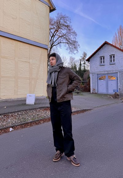 Chaqueta de cuero marrón, camisa negra, pantalones negros, bufanda gris. Zapatos marrones con suelas blancas. Configuración casual al aire libre con edificios y árboles.