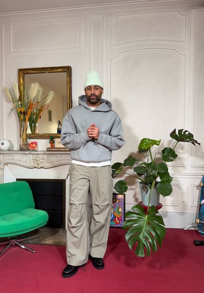 Sudadera con capucha gris, pantalones cargo beige, zapatos negros. Entorno interior con silla verde, planta en maceta y espejo decorativo en el fondo.