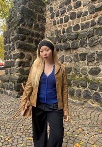 Tan leather jacket over a blue ribbed cardigan, black wide-leg trousers, and a black headband. The background includes a stone wall and a cobblestone path.