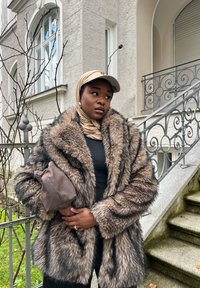 Barna műszőrme kabát hosszú, puha szálakkal és fekete csíkokkal, fekete felső fölött viselve. A modell barna kézitáskát tart, és bézs sapkát valamint sálat visel.