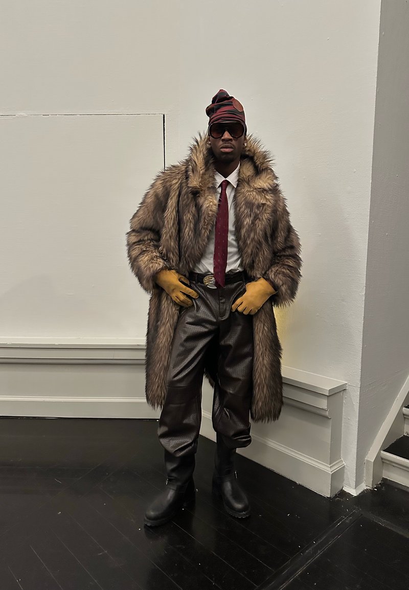 Homme portant un manteau en fourrure surdimensionné, chemise blanche, cravate rouge, gants jaunes, pantalon en cuir, bottes noires, lunettes de soleil et un bonnet rouge et noir à motifs à l'intérieur.