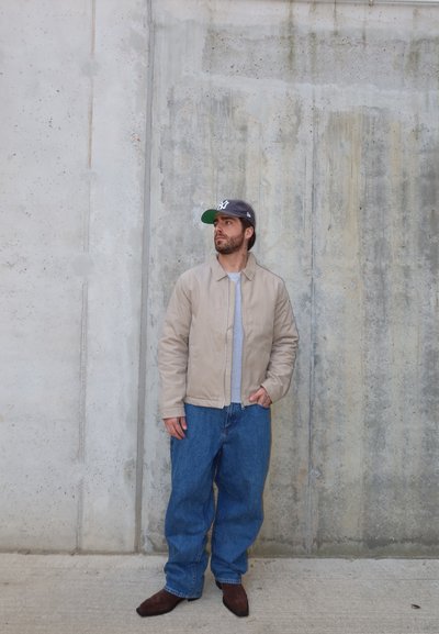 Hombre con chaqueta beige, vaqueros azules, zapatos marrones y gorra negra de pie contra una pared de hormigón texturizada, mirando hacia un lado.
