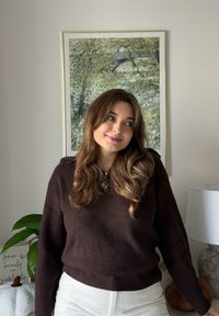 Ciemnobrązowy prążkowany sweter z dekoltem w serek, zestawiony z białymi spodniami z wysokim stanem. W tle zielone liście na obrazie oraz lampa.