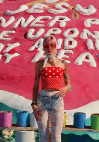 Rdeč zgornji del obleke s pikami, bele legice z čipko, s šal na glavi. Barvne vedra s barvami v ozadju ob rožnati teksturirani steni.