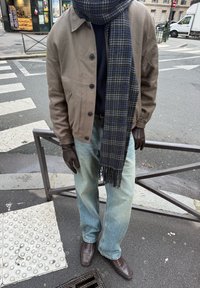 Άτομο που φοράει μπεζ μπουφάν, μπλε καρό κασκόλ, μπλε τζιν, καφέ δερμάτινα γάντια και καφέ παπούτσια, στέκεται σε πεζοδρόμιο πόλης κοντά σε διάβαση πεζών.