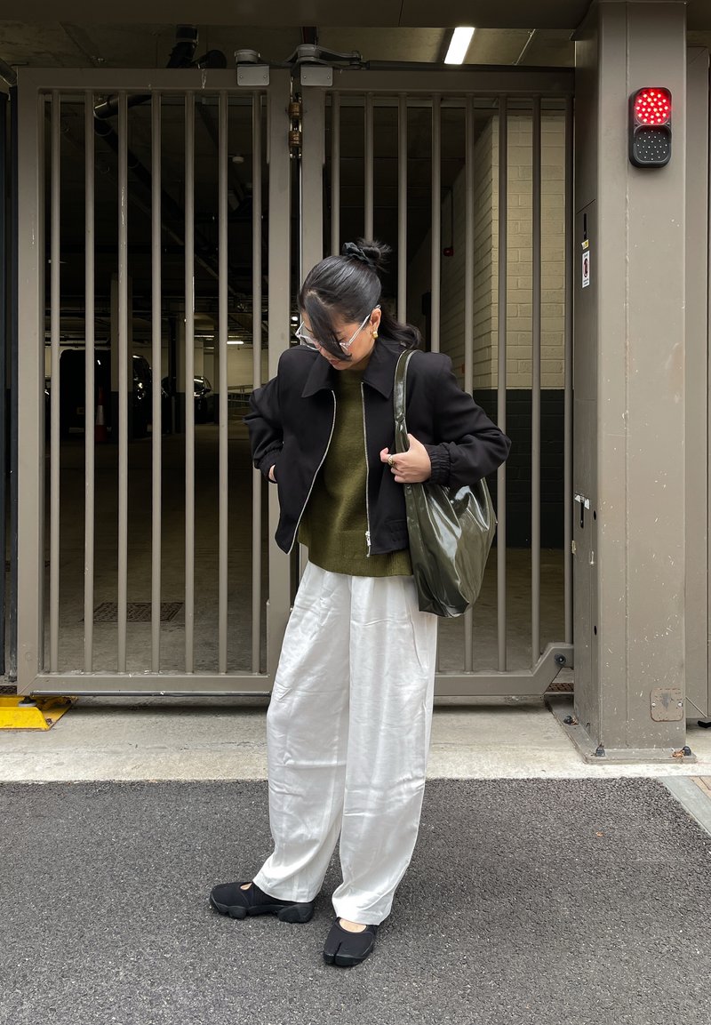 Veste noire zippée, pull vert olive, pantalon blanc à jambes larges, et chaussures noires avec un design unique au niveau des orteils. Tenant un sac fourre-tout olive brillant.