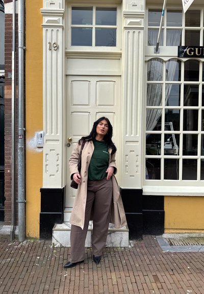 Abrigo trench beige sobre una camisa verde, combinado con pantalones anchos marrones. De pie sobre un pavimento de ladrillo frente a un edificio amarillo con puerta blanca.