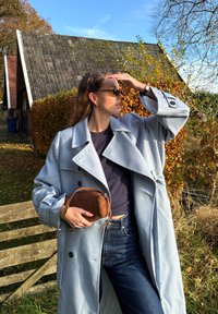 Jasnoniebieski oversize płaszcz z dużymi klapami, czarne guziki; ciemna bluzka, dżinsy, brązowa zamszowa torebka. Na zewnątrz, w tle jesienne liście.