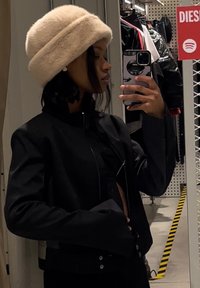 Μαύρο κοντό μπουφάν με ψηλό γιακά και κουμπιά που κλείνουν με κουμπί, συνδυασμένο με ένα ανοιχτό μπεζ καπέ�λο από γούνα. Ανάκλαση σε καθρέφτη με φόντο κατάστημα.