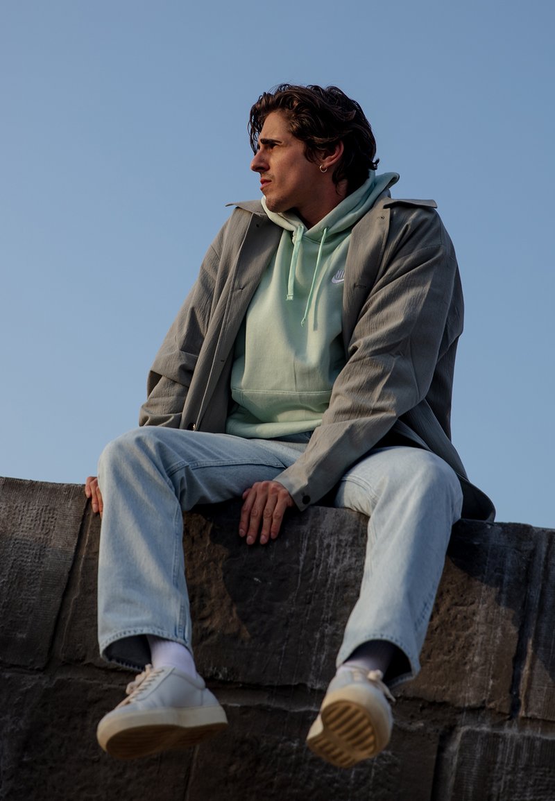 Neformalus apranga, kurioje matomas šviesiai pilkas paltas uždėtas ant mėtų žalios spalvos džemperio su gobtuvu, šviesiai mėlyni džinsai ir baltos sportinės bateliai su tekstūruotu padu, sėdintis ant akmeninio borto.