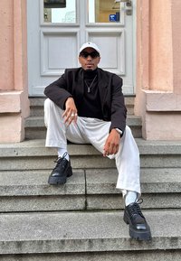 Scarpe platform nere chunky, calzini bianchi, pantaloni bianchi larghi, e un dolcevita nero sotto una giacca nera, accessoriati con una collana e occhiali da sole.