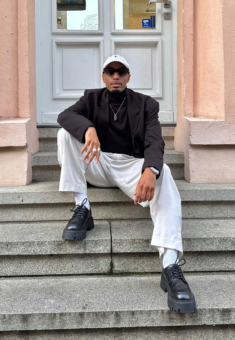 Chaussures plateformes noires épaisses, chaussettes blanches, pantalons blancs amples et un col roulé noir sous un blazer noir, accessoirisés d'un collier et de lunettes de soleil.