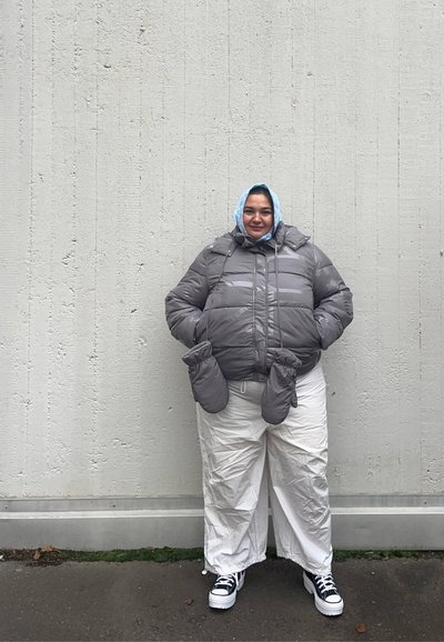 Chaqueta de abrigo gris con capucha y guantes integrados, combinada con pantalones de pierna ancha blancos. Zapatillas de plataforma en blanco y negro completan el atuendo.