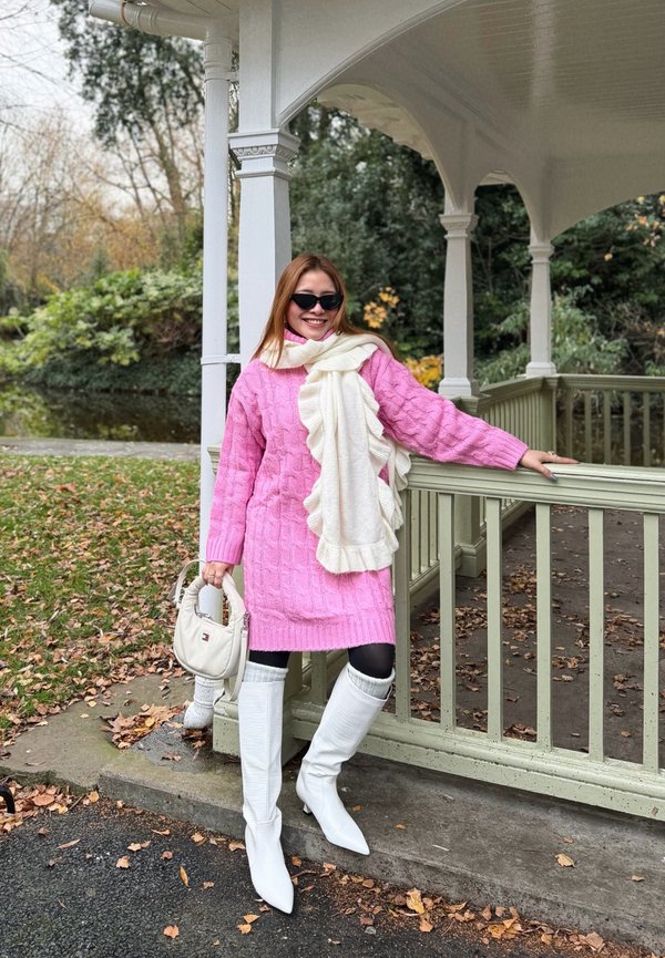 Donna con vestito rosa a trecce, sciarpa bianca arricciata, stivali bianchi e occhiali da sole, in piedi accanto alla ringhiera di un padiglione di legno verde pallido all'aperto.