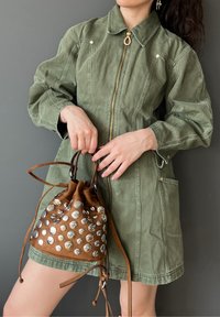 Robe en denim vert olive avec fermeture éclair à l'avant, détails à pression et deux poches latérales. Le modèle tient un sac à cordon marron avec des clous métalliques.