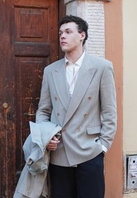 Lys grå dobbeltradet blazer med en struktureret finish, parret med en hvid skjorte og mørke bukser. En lys frakke holdes i den ene hånd.