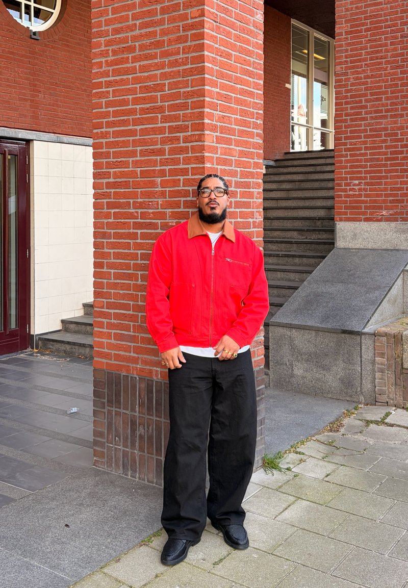 Homme aux cheveux tressés et lunettes, portant une veste rouge et un pantalon noir, debout contre un pilier en briques rouges près des escaliers.