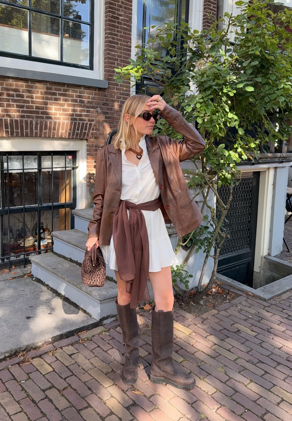 Bruine leren blazer over een witte jurk met een bruine ceintuur, kniehoge bruine laarzen en een geweven bruine handtas, opgesteld op een straat van bakstenen.