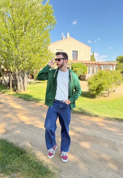 Sudadera verde con cremallera, camiseta blanca, jeans azules y zapatillas rojas. De pie en un camino de tierra con vegetación y una casa de fondo.