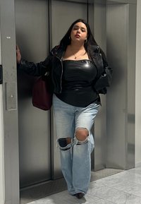 Una donna con giacca di pelle nera e top, indossa jeans chiari strappati e si trova davanti a porte dell'ascensore chiuse mentre tiene in mano una borsa di colore rosso scuro.