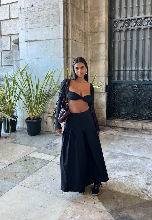 Femme portant un crop top noir et une longue jupe, debout dans un couloir en pierre près de plantes en pot et d'une grande grille en fer.