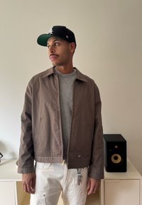 Ruskea vetoketjullinen takki kauluksella, puettu harmaan neuleen päälle. Takissa on sileä pinta ja kaksi sivutaskua.