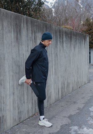 Chaqueta deportiva negra con cuello alto, combinada con leggings negros ajustados a rayas y zapatillas de running blancas, frente a una pared de concreto.