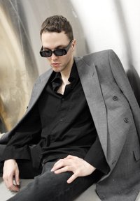 Blazer gris croisé posé sur une chemise noire à boutons et un pantalon foncé, porté avec des lunettes de soleil noires angulaires, assis devant un fond métallique.