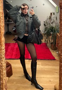 Ελαιόχρωμο bomber jacket, καφέ cropped top, μαύρες βερμούδες, διάφανες καλσόν, μαύρες δερμάτινες μπότες μέχρι το γόνατο και μια μαύρη τσάντα ώμου. Αντανάκλαση στον καθρέφτη.