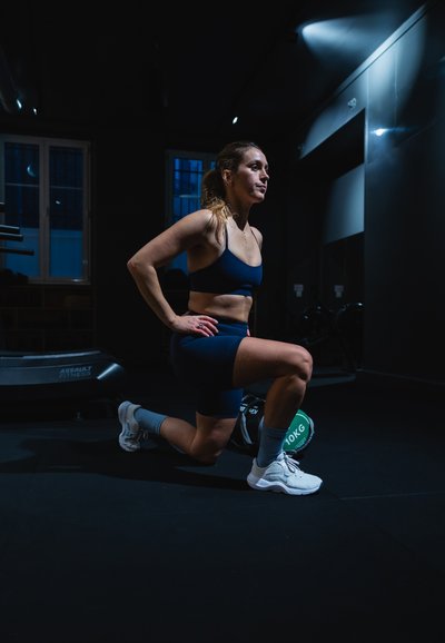 Mujer atlética en un gimnasio oscuro, vistiendo un crop top azul marino y shorts, realizando una zancada al lado de un peso verde de 10 kg, con zapatillas grises.