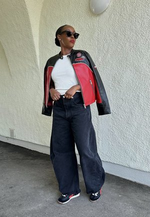 Schwarze und rote Lederjacke mit hohem Kragen, weißes Oberteil, übergroße schwarze Jeans und schwarze Sneakers mit roten Akzenten; stilvolle Sonnenbrillen runden den Look ab.