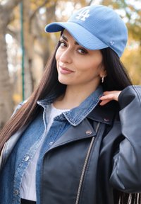 Gaismas zilas krāsas cepure ar auklu un dekoratīvu logo priekšpusē, kombinēta ar slāņotu džinsa jaku. Melni ādas elementi un balta topiņa noslēdz izskatu.